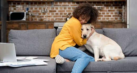 Vrouw in gele trui die op een grijze bank zit en haar blonde labrador knuffelt.